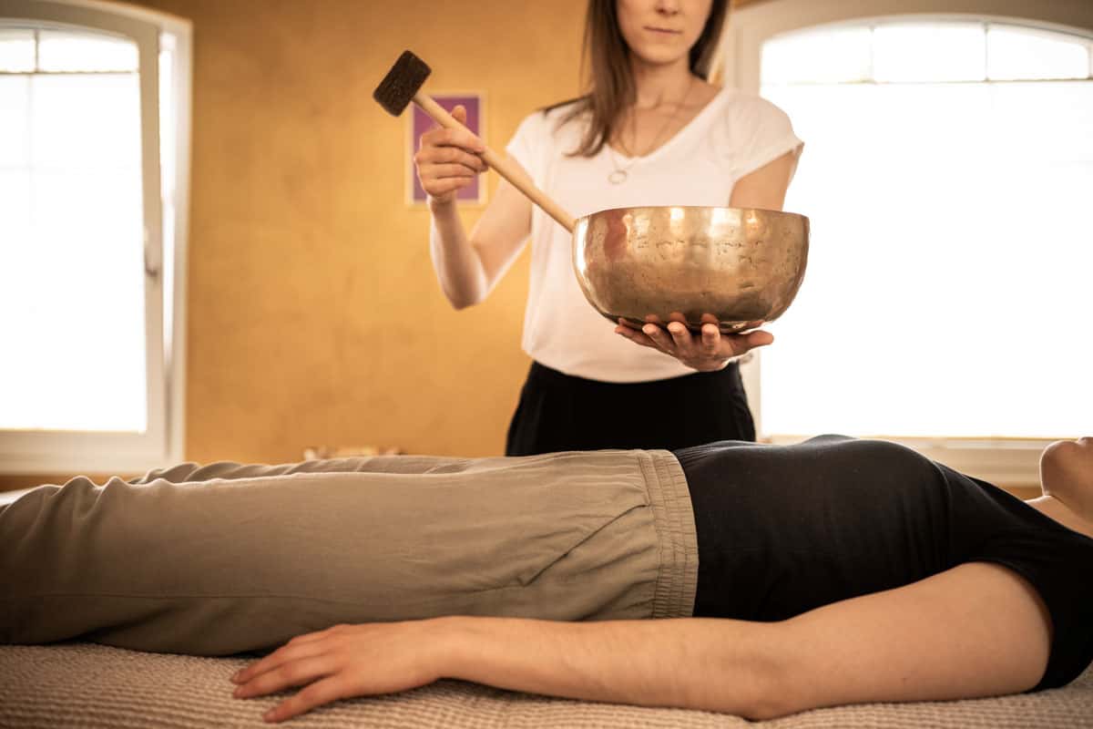 Eden Bratoljic schlägt mit dem Klöppel gegen Klangschalen die sie in der Hand hält. Darunter liegt eine Klientin.